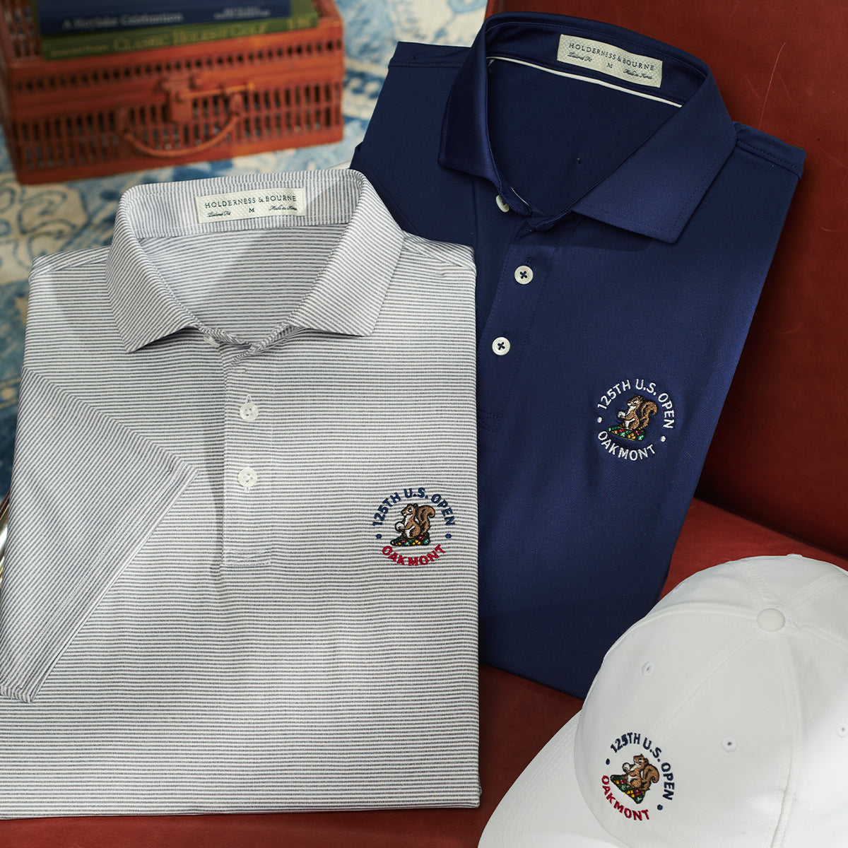 Folded gray striped and navy polos and a white cap, each embroidered with a "125TH U.S. OPEN OAKMONT" crest with a squirrel.