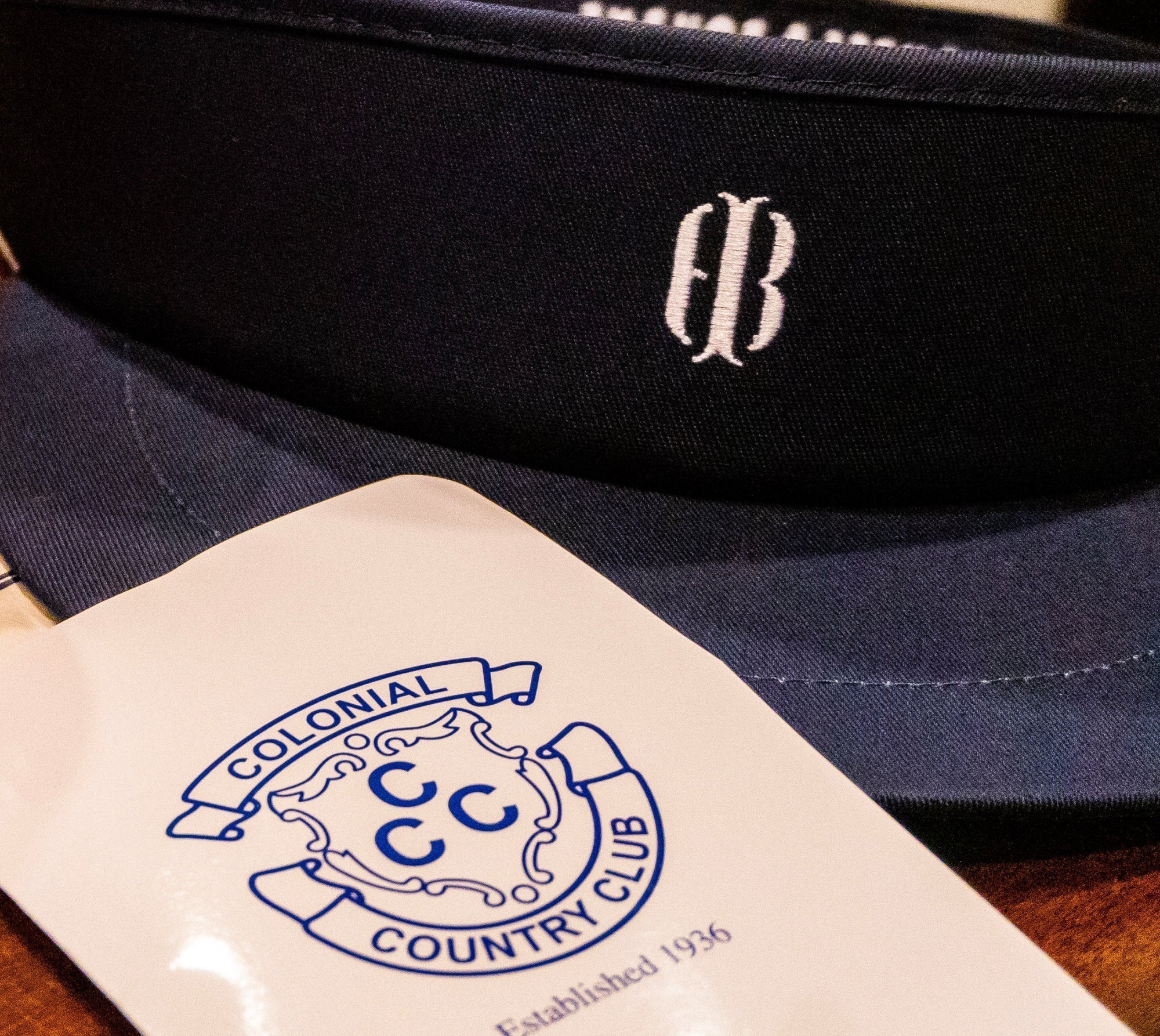Navy visor with a white embroidered monogram, resting beside a card tag from Colonial Country Club, Established 1936.