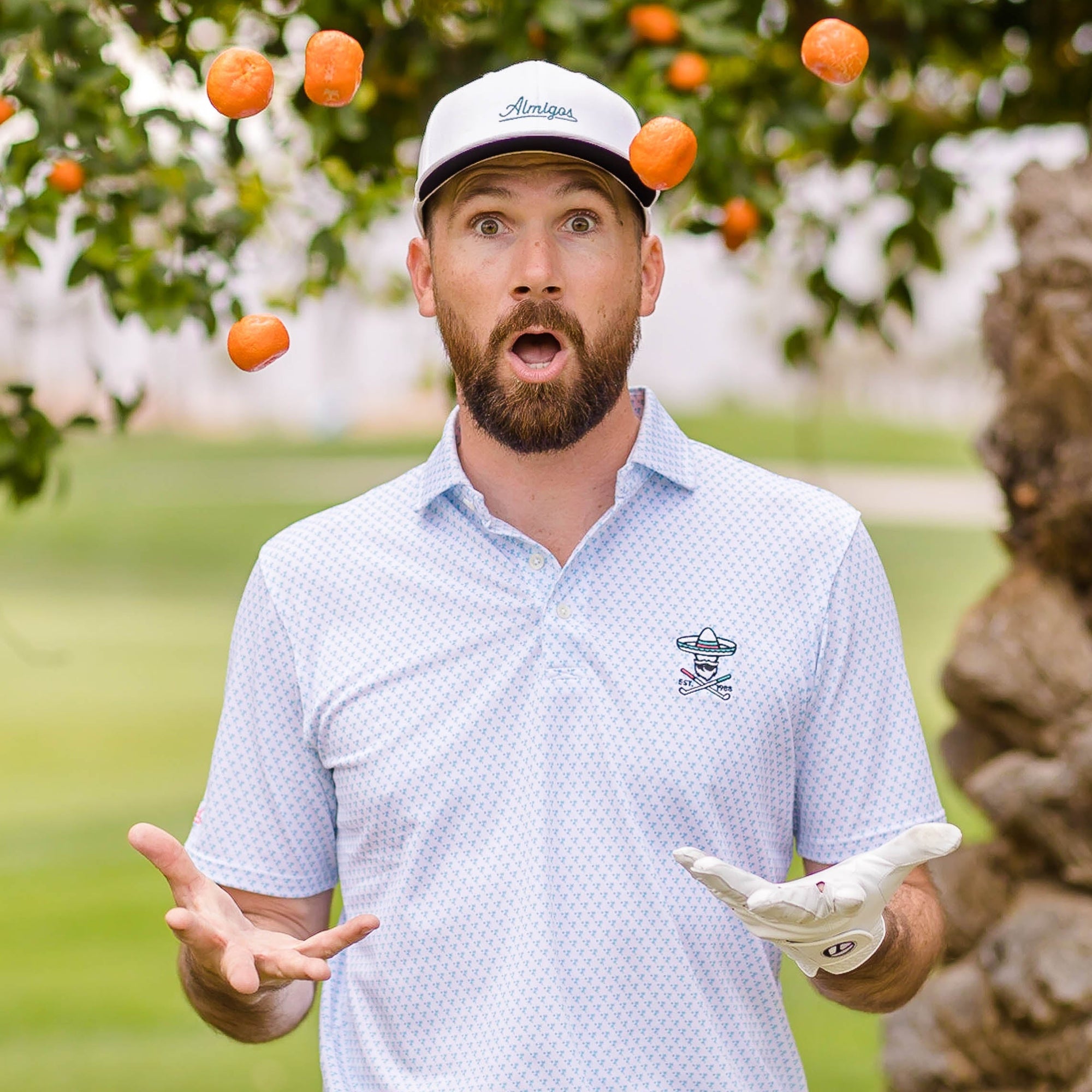 Surprised bearded man in a white "Almigos" cap and light blue patterned polo with oranges tossed in the air around him.