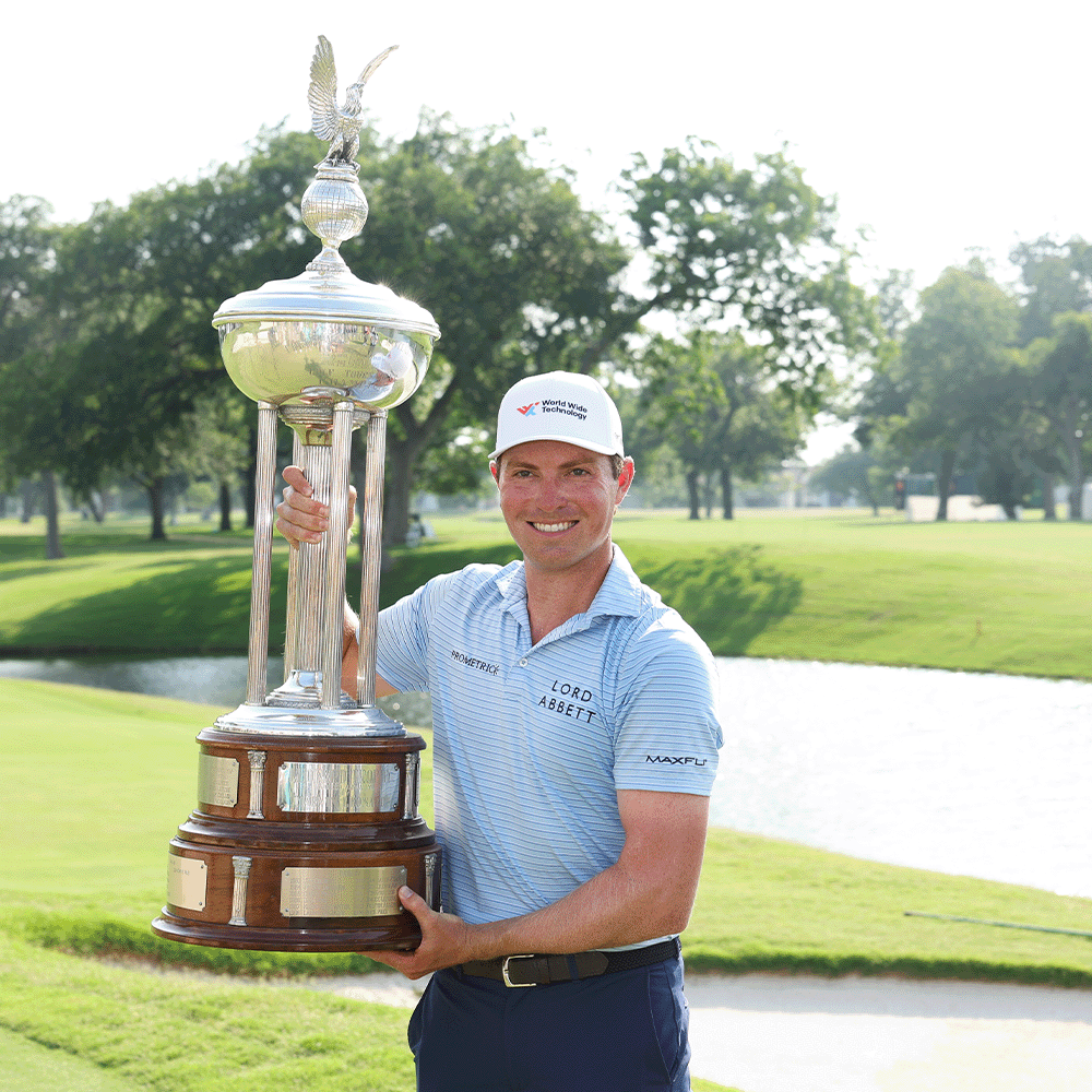 Smiling golfer in a white cap and light blue striped polo holding a large silver trophy on a golf course beside a pond.