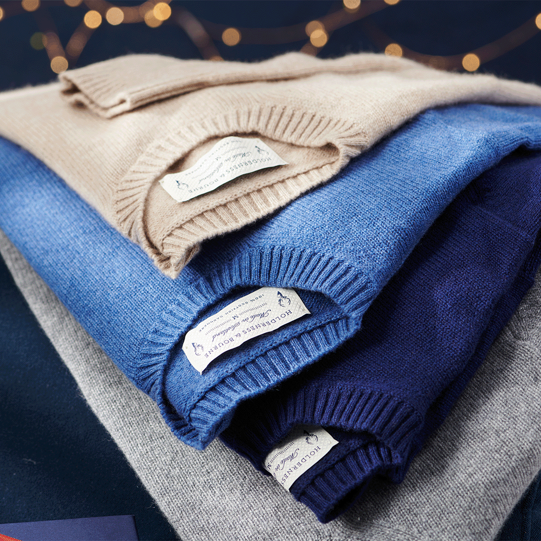 Stack of folded sweaters in tan, blue, navy and gray with Holderness & Bourne neck labels and holiday string lights behind them.