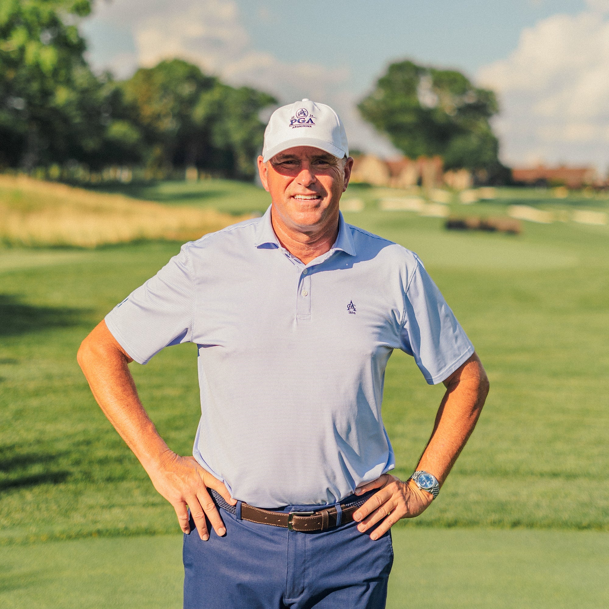 Smiling man in a white PGA cap, light blue polo and navy pants standing with hands on hips on a golf course.