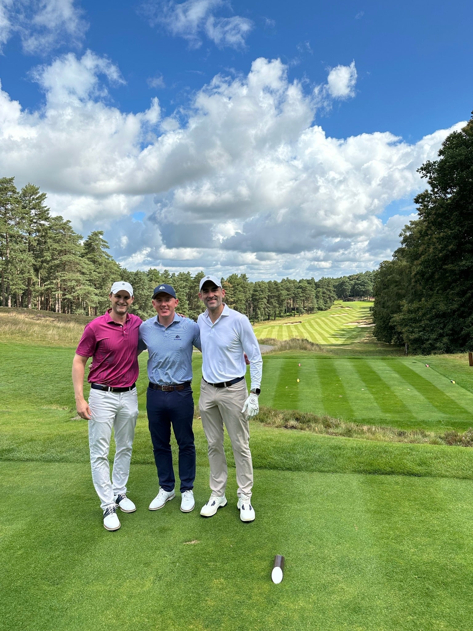 Three men in golf clothes smiling arm in arm on a tee box, with a striped fairway and pine trees behind them.