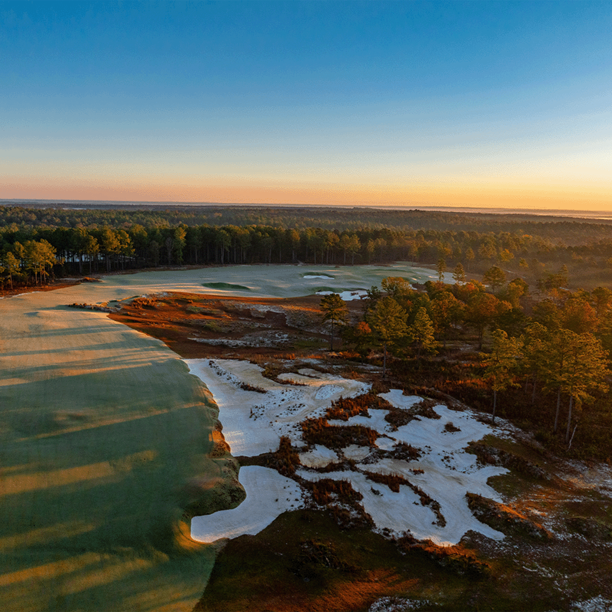 Aerial view of a golf course fairway and white sand bunkers under an orange horizon glow, with pine trees in autumn color.