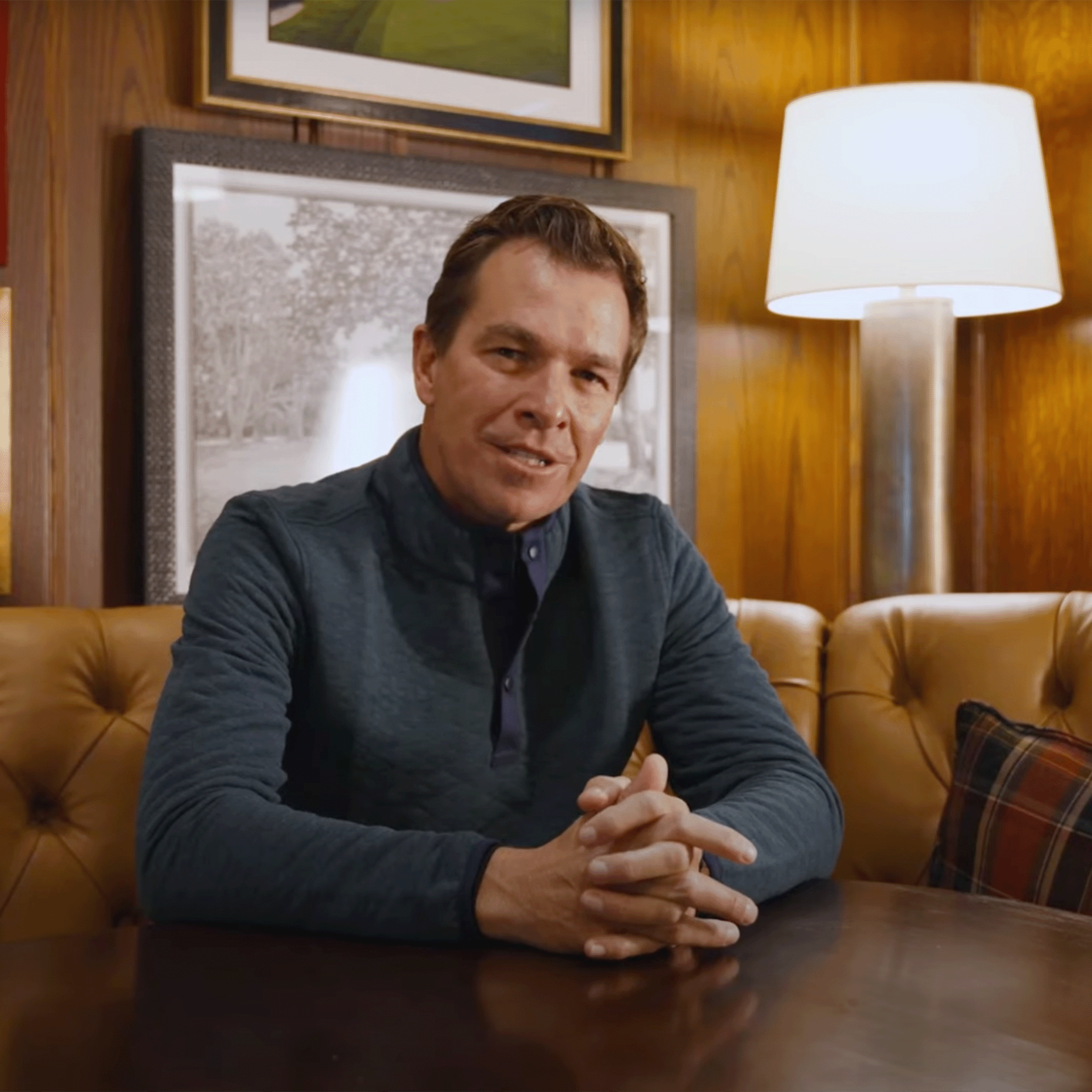 Man in a dark quilted quarter-zip seated at a wooden table with hands clasped, in a paneled room with a tufted leather sofa.