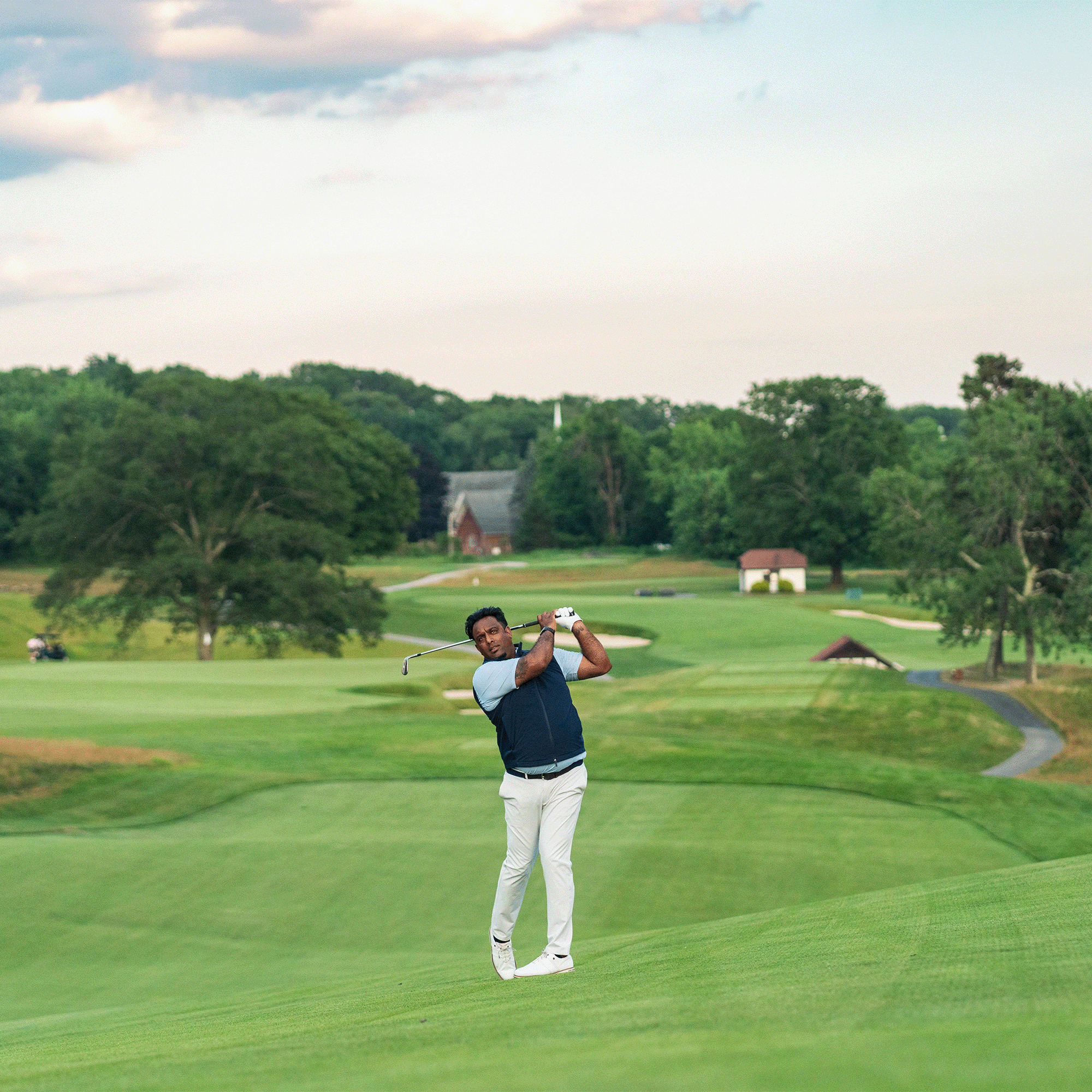 Golfer in a navy vest, light polo and white pants finishing a swing on a rolling course with a barn behind him.