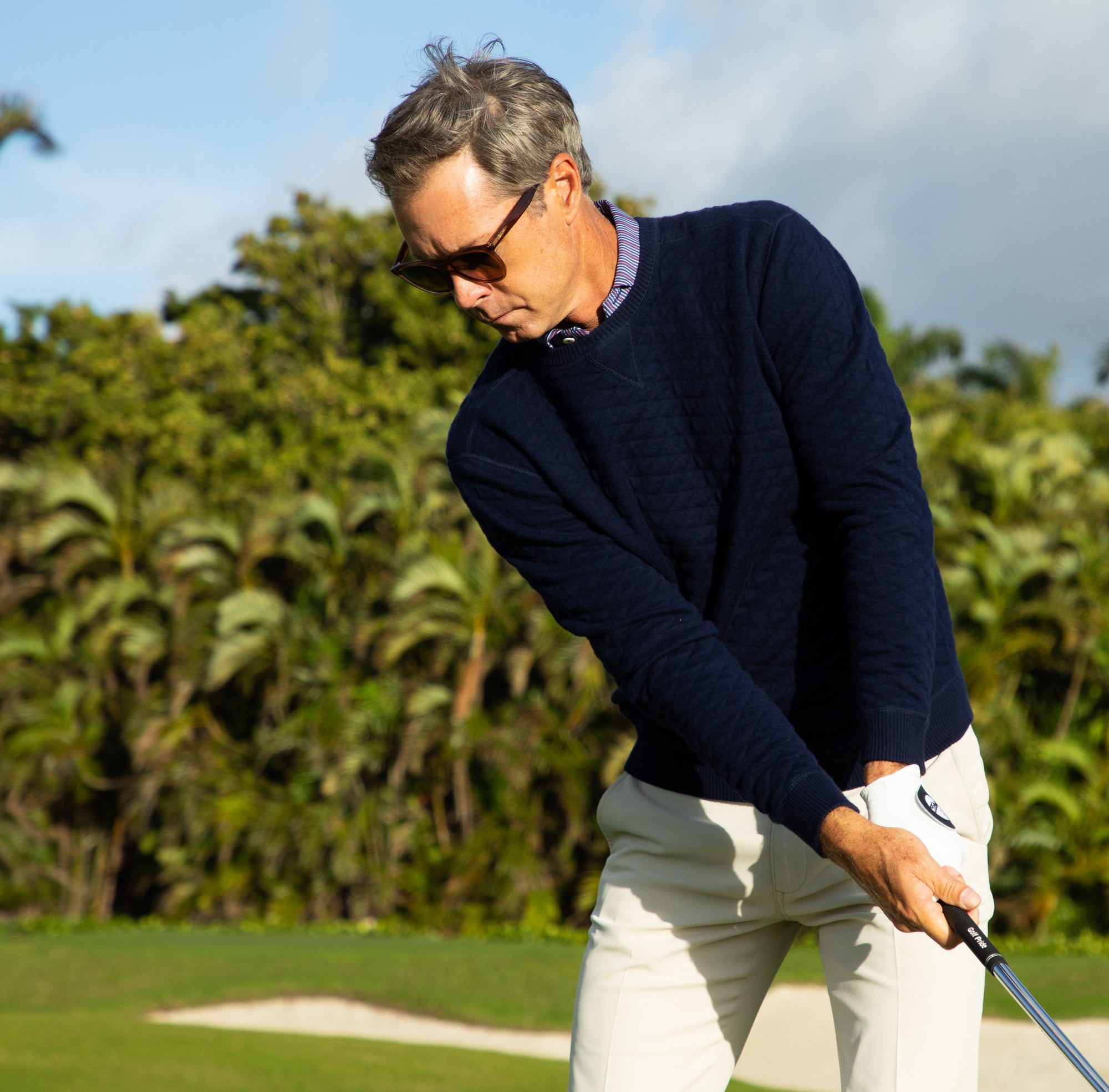 Golfer in sunglasses, a navy quilted sweater and stone pants gripping an iron in his gloved hand on a tropical course.