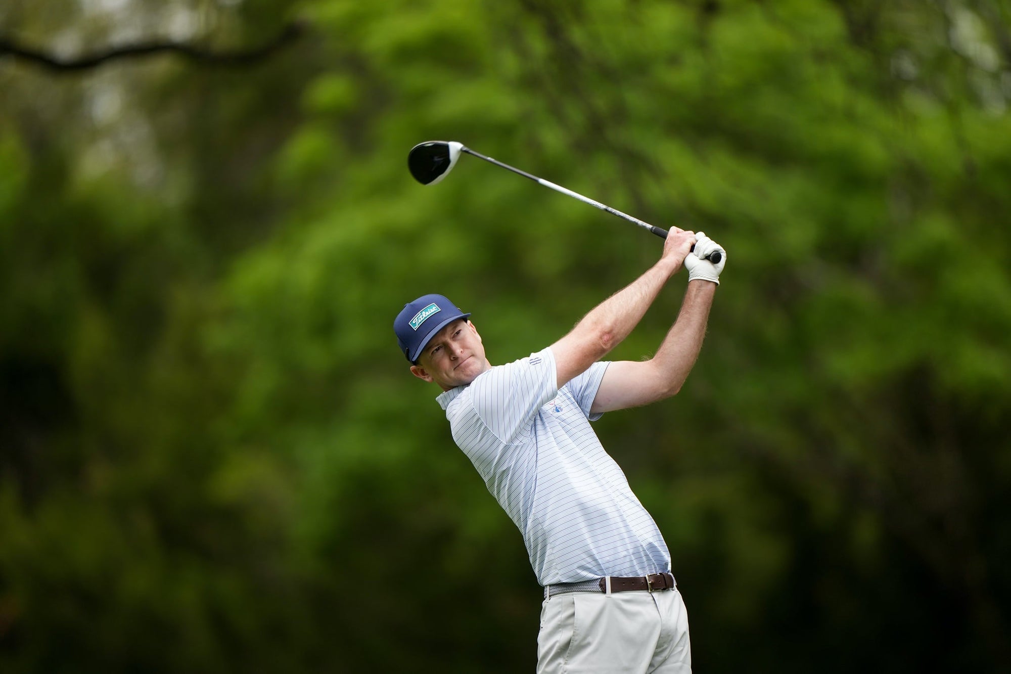 Golfer in a navy cap, light blue striped polo and brown belt following through on a drive against green trees.