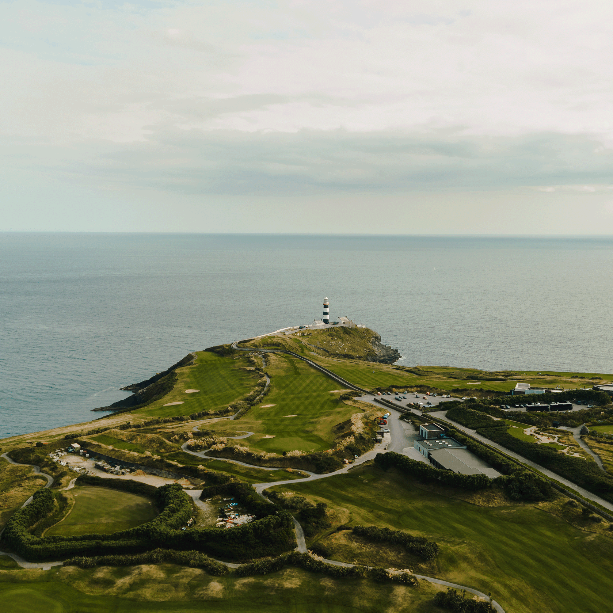 Aerial view of a coastal golf course on a headland, with a black and white striped lighthouse at the tip and the sea beyond.