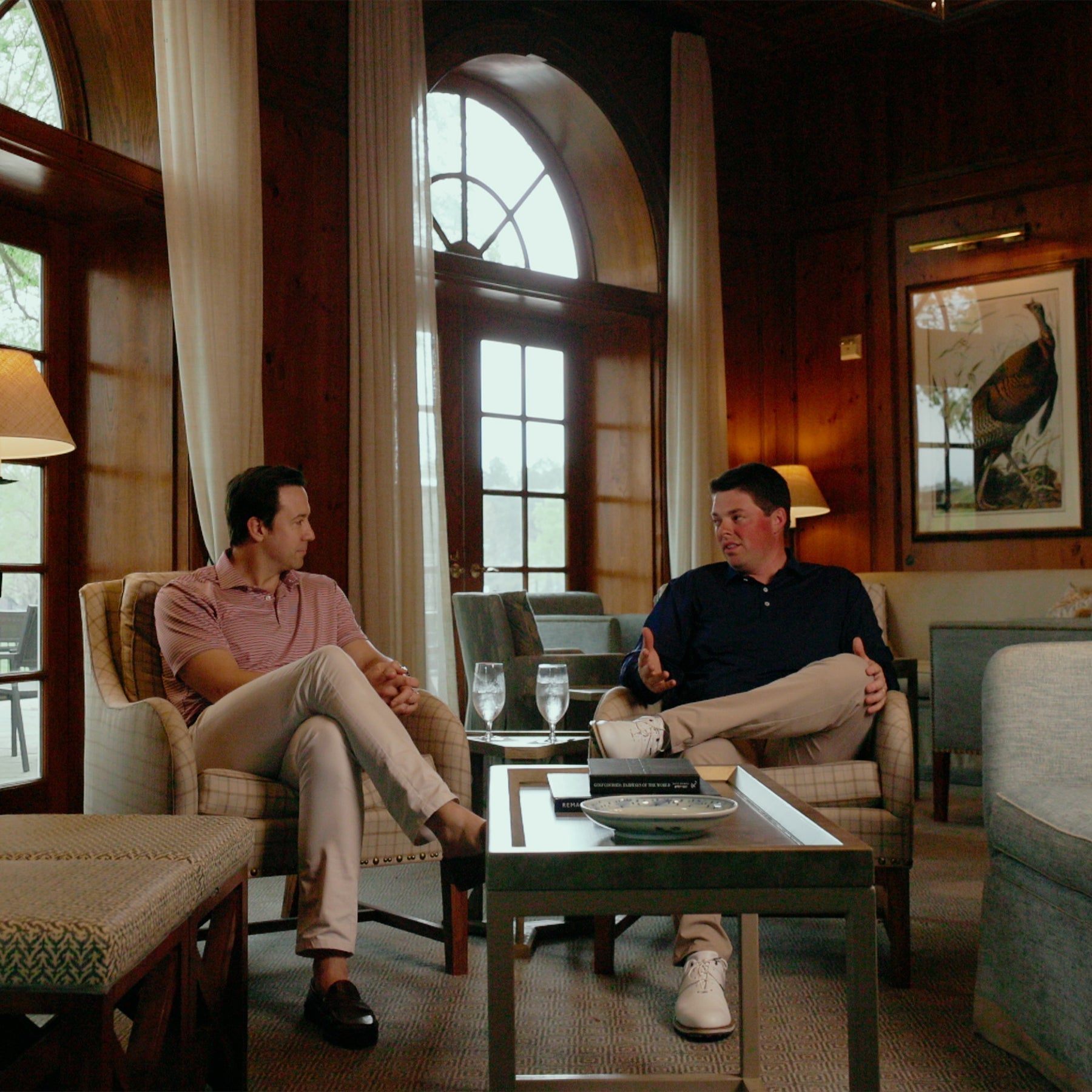 Two men in conversation in wood-paneled club lounge chairs, one in a pink striped polo and one in a navy shirt.