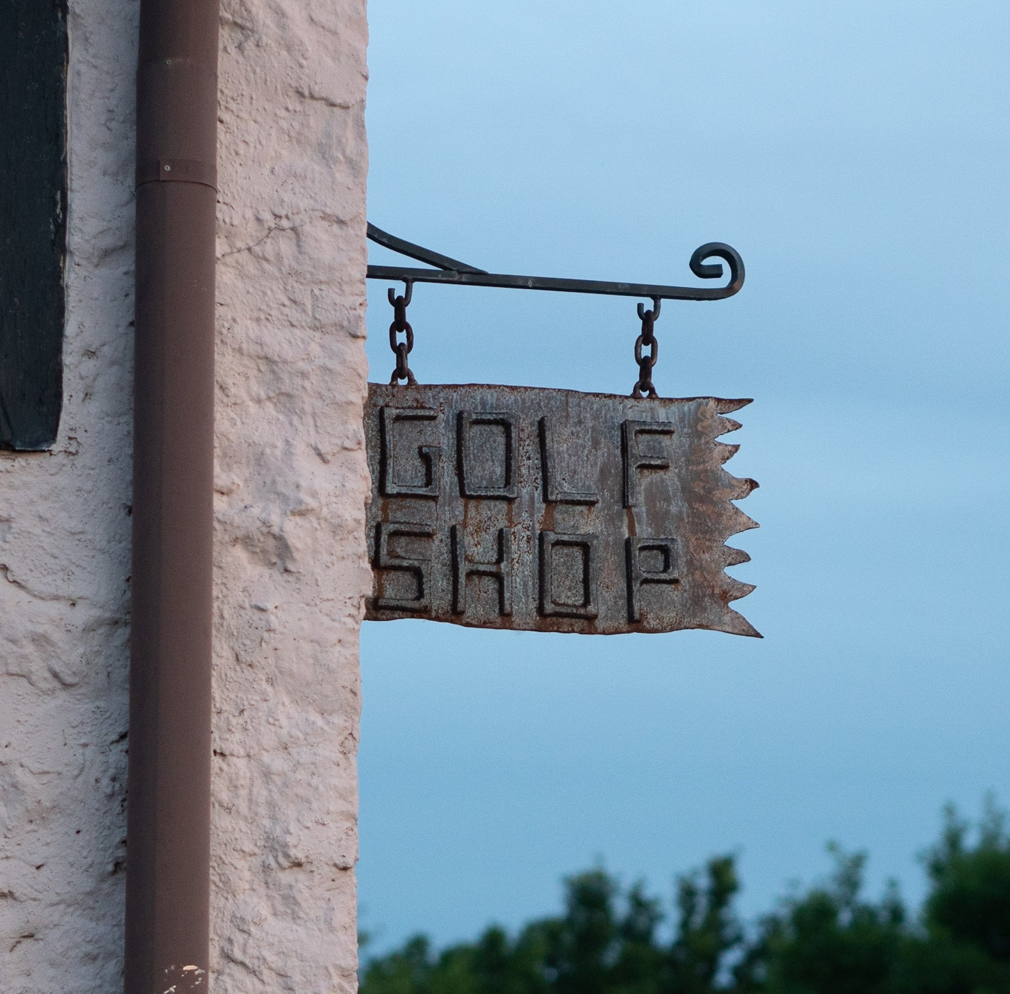 Rusty hanging iron sign reading "GOLF SHOP" on a pale stucco wall against a dusky blue sky.