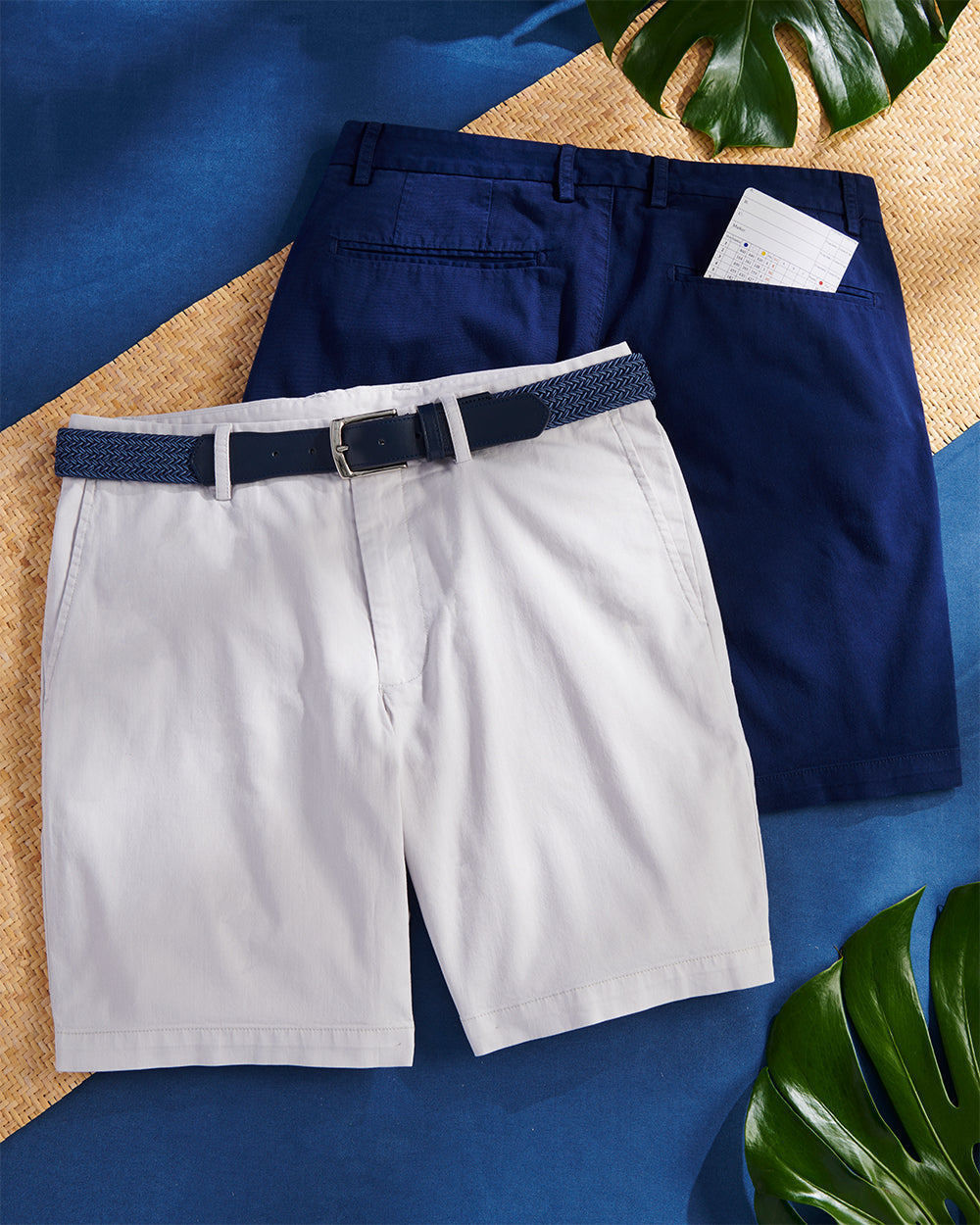 Light gray and navy shorts laid flat with a woven navy belt, on a blue backdrop with palm leaves and a scorecard.