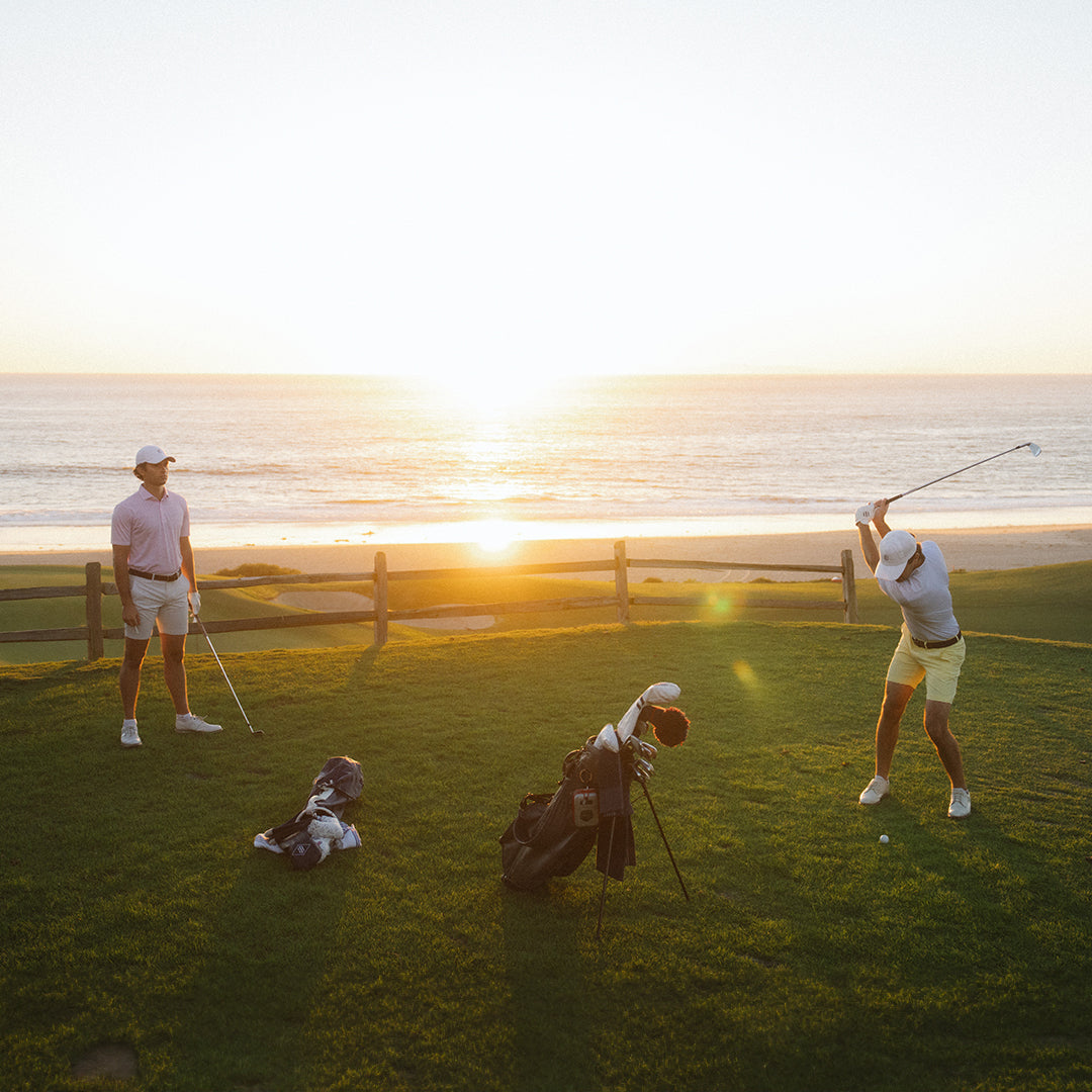 Two golfers on a seaside course at sunset, one in a pink polo watching as the other swings in a light polo and yellow shorts.