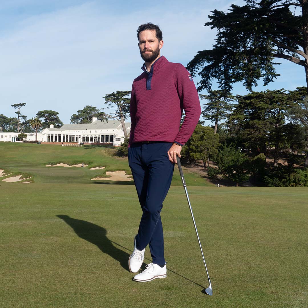 Bearded golfer in a maroon quilted quarter-zip and navy pants leaning on an iron, with a white clubhouse and cypress trees behind him.