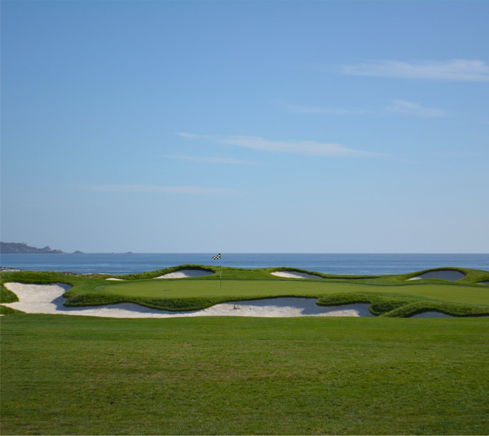 Seaside golf green with a checkered flag, white sand bunkers and the ocean under a pale blue sky.