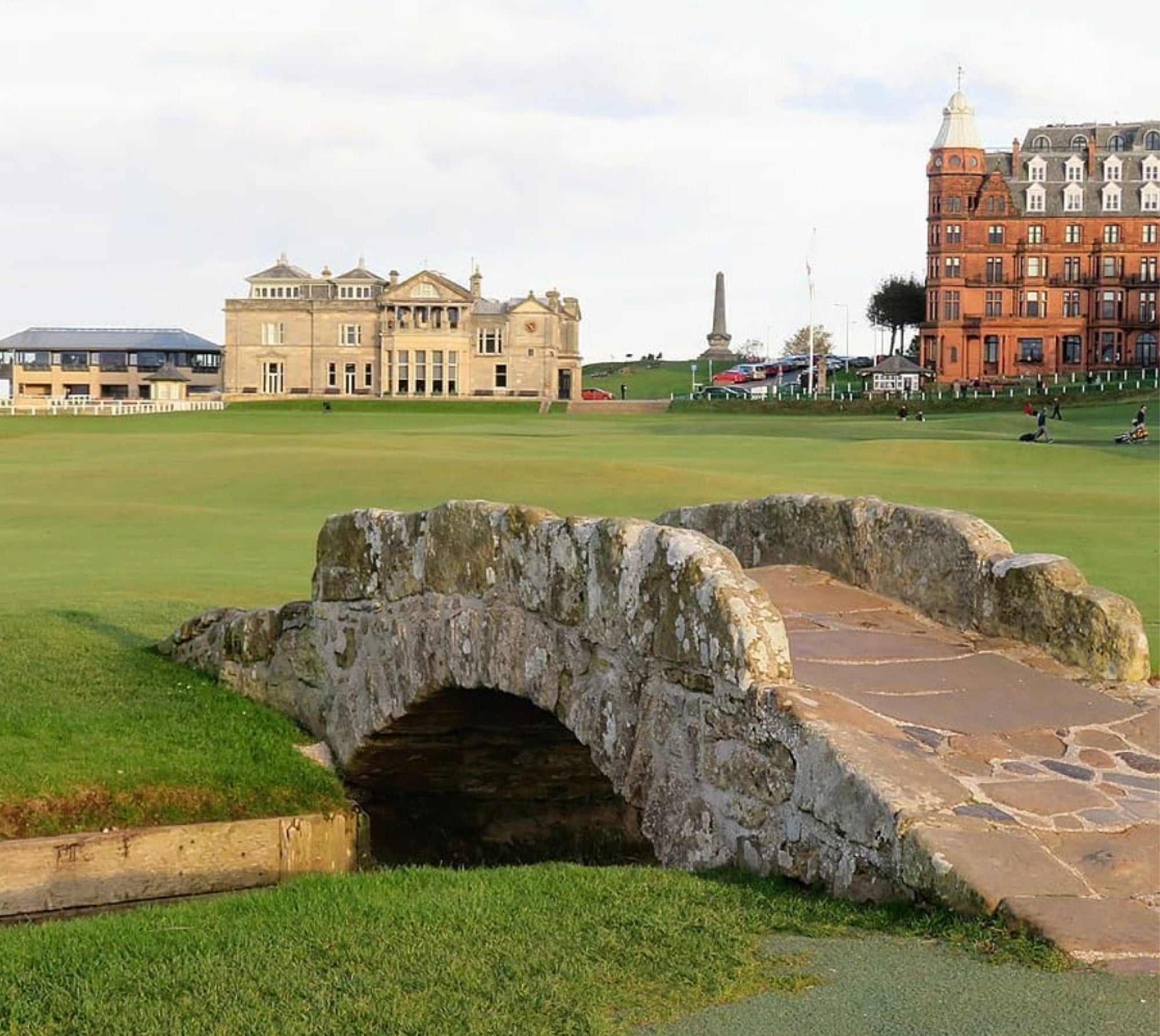 Old stone bridge over a burn on a links fairway, with a stately clubhouse and a red brick hotel behind.