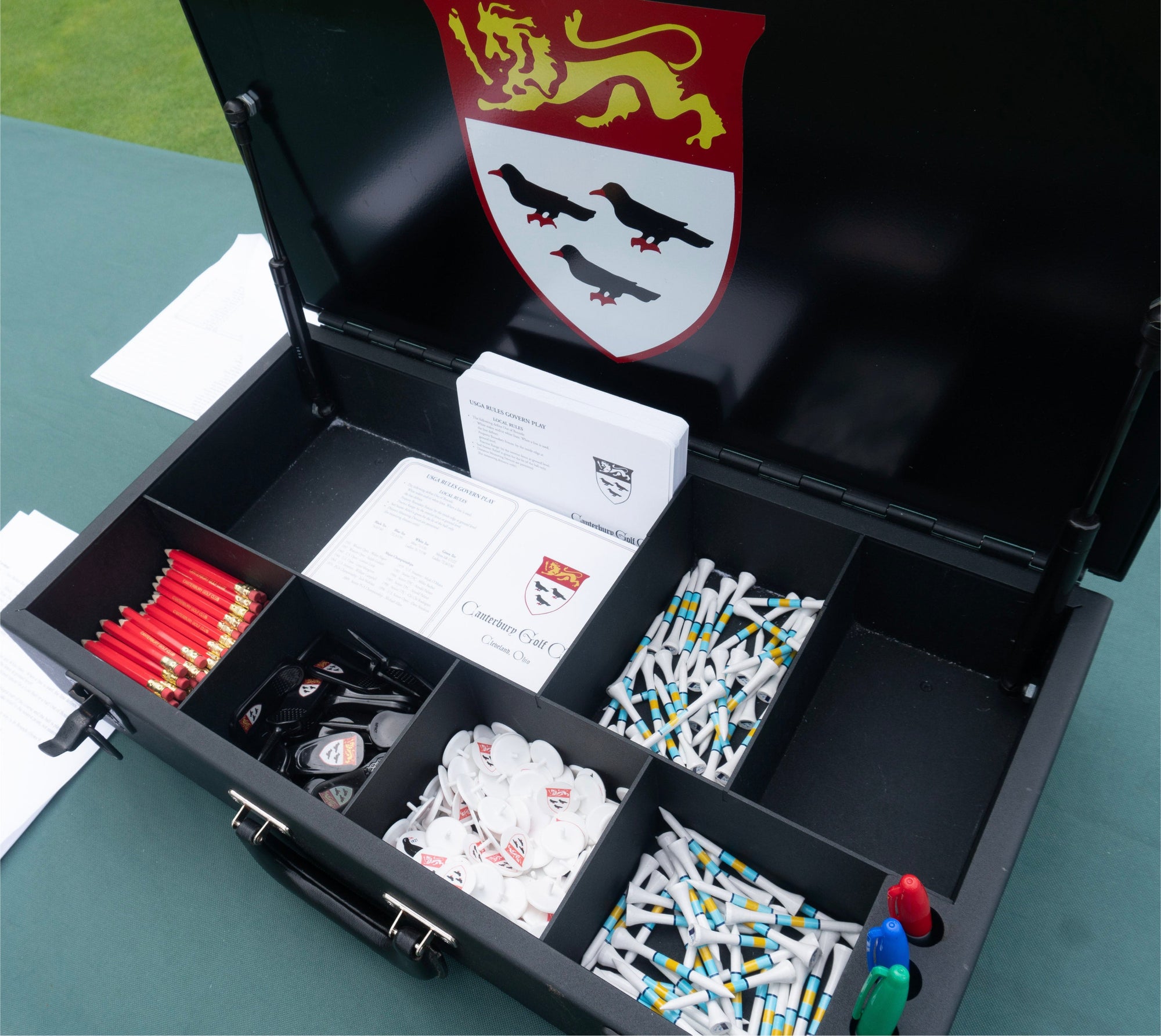 Open black case of golf supplies with red pencils, tees, ball markers, scorecards and a crest on the lid.