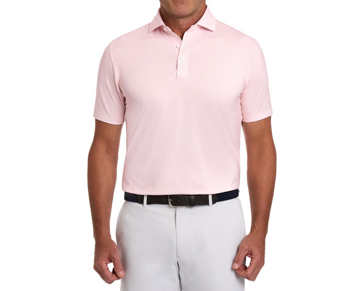 The Hanover Shirt: Nectarine, a short-sleeve polo with geometric jet pattern, tucked into light-gray trousers, featuring a dark woven belt.