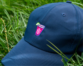 Navy golf hat with embroidered Holderness and Bourne pink cocktail logo in patch of grass.