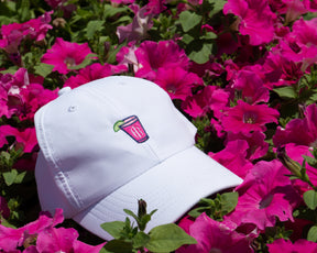 Holderness and Bourne white performance hat with branded pink cocktail logo perched on top of a bush with pink flowers.