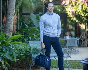 Man holding duffel wears a grey sweater in a park with benches.