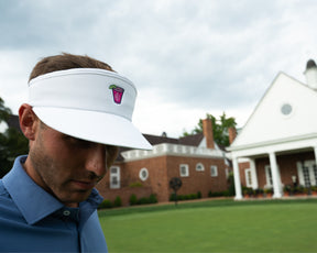 Male golfer wearing Holderness and Bourne white visor stands in front of green lawn and brick building.