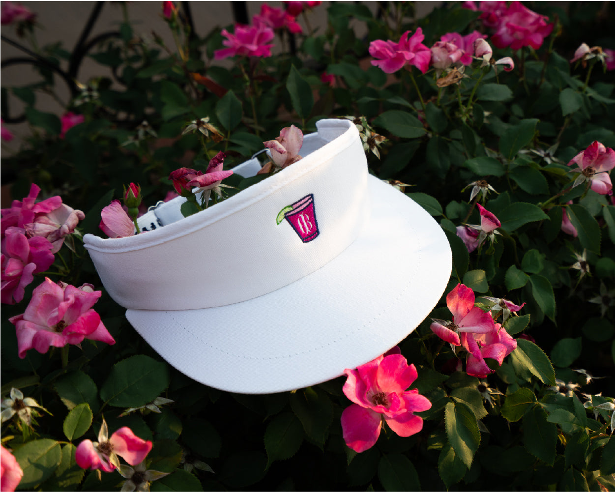 Holderness and Bourne white tour visor golf hat with branded pink cocktail logo perched on top of a bush with pink flowers.