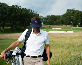 Male golfer wearing navy Holderness and Bourne visor for golf carries golf club bag and walks across golf course.
