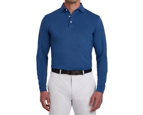 The Abbott Shirt: Andover, a long-sleeve polo in blue, worn with gray trousers. Features a spread collar, contrast piping, and breathable performance fabric.