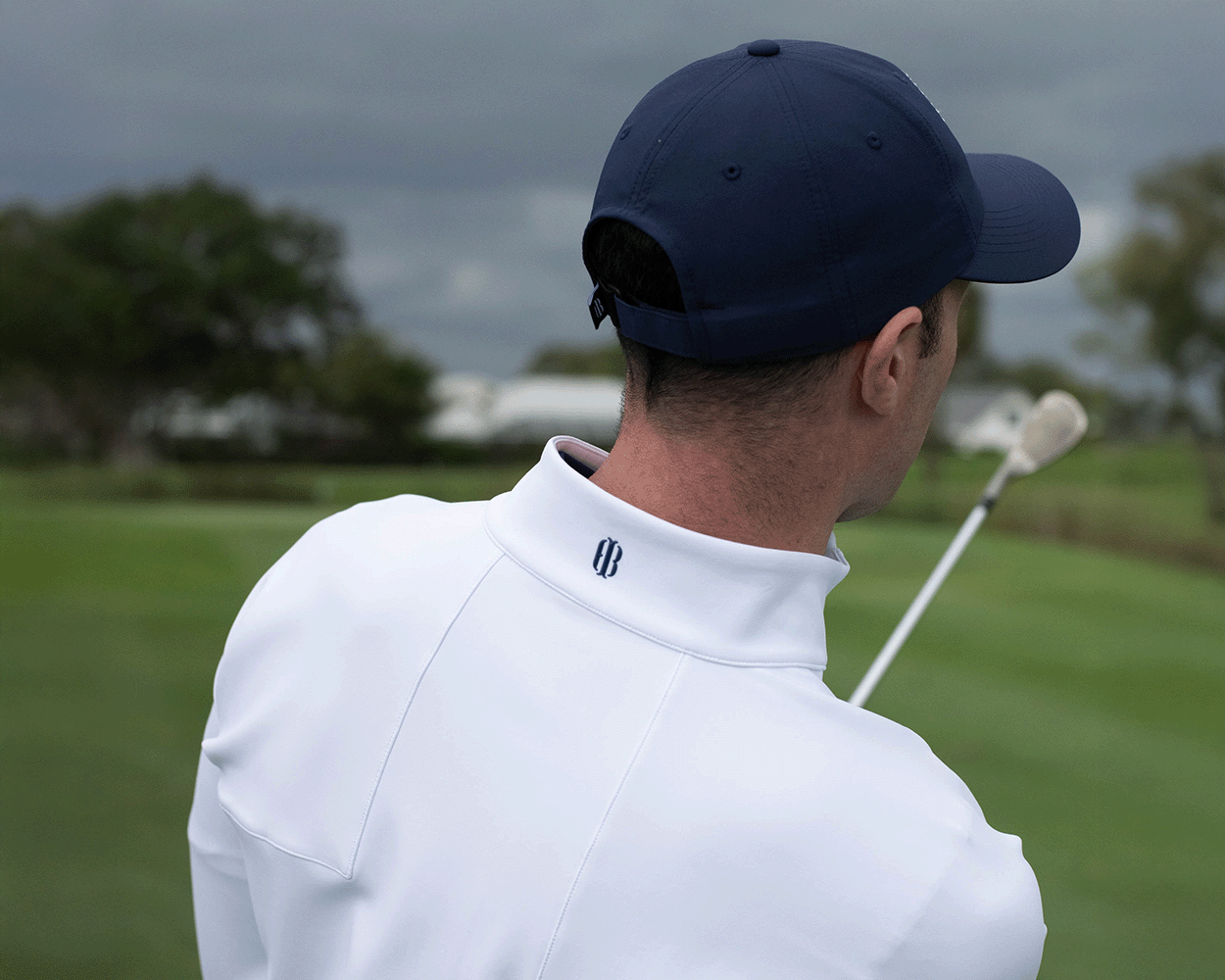 Man on golf course wearing 2026 U.S. Open Rodgers Pullover: White, holding golf club over shoulder, showcasing pullover's high collar and logo detail.