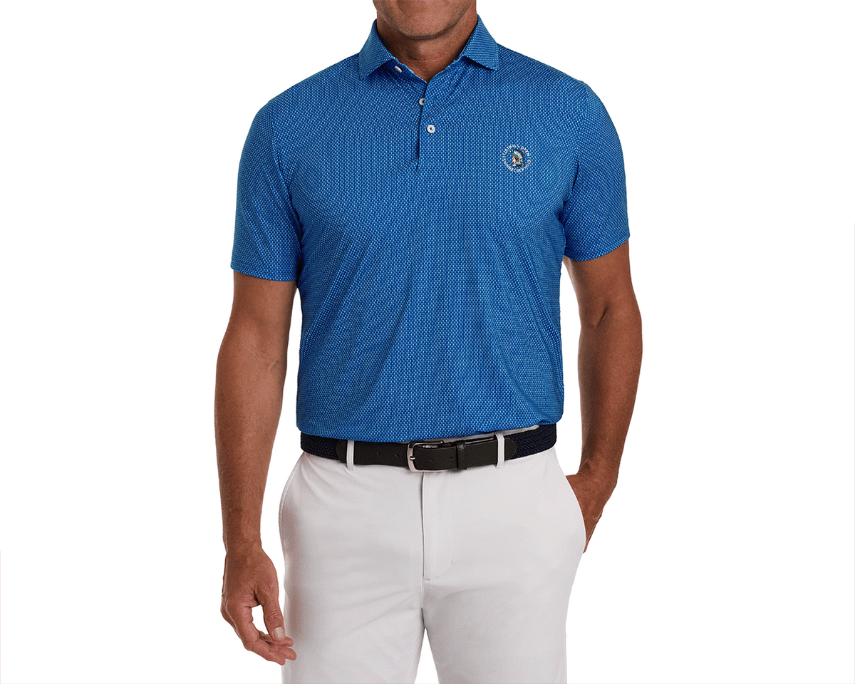 Man wearing the 2026 U.S. Open Hicks Shirt: Maidstone Blue, featuring a geometric dot print, embroidered logo, white trousers, and a dark woven belt.