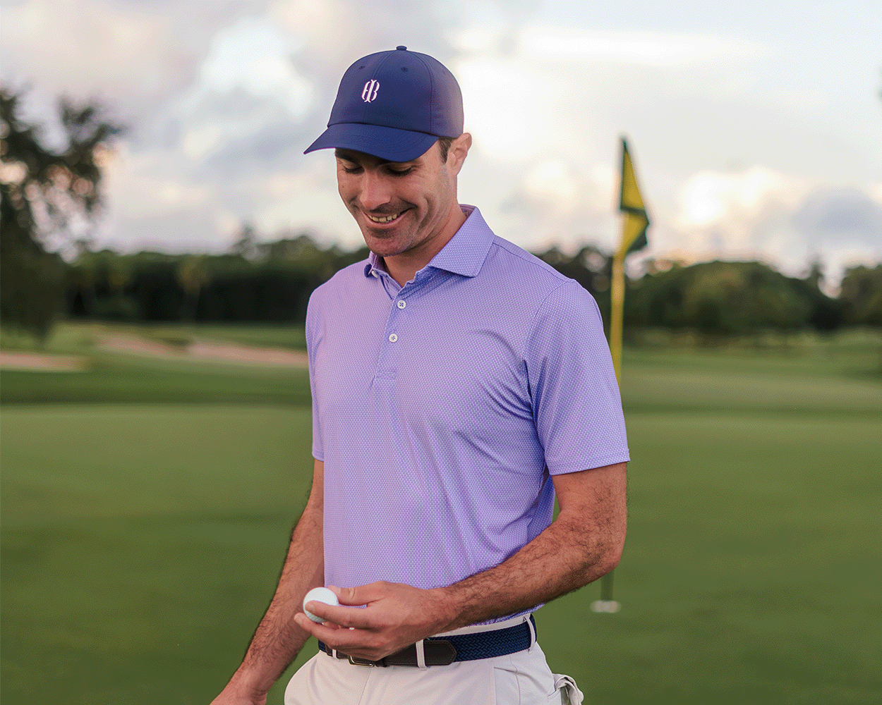 Man on golf course wearing The Hicks Shirt: Tudor, a geometric-pattern polo, holding a golf ball with flagstick and greenscape in the background.