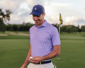 Man on golf course wearing The Hicks Shirt: Tudor, a geometric-pattern polo, holding a golf ball with flagstick and greenscape in the background.