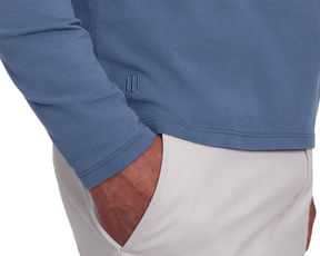 Detail of The Montague Long Sleeve T-Shirt: Pacific Blue showing the small embroidered logo mark at the lower hem, with a model's hand in a light gray pocket.