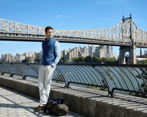A young man wears The Swenson Vest: Andover, a quilted vest ideal for warmth and breathability, leaning on a riverside railing with a duffel bag nearby.