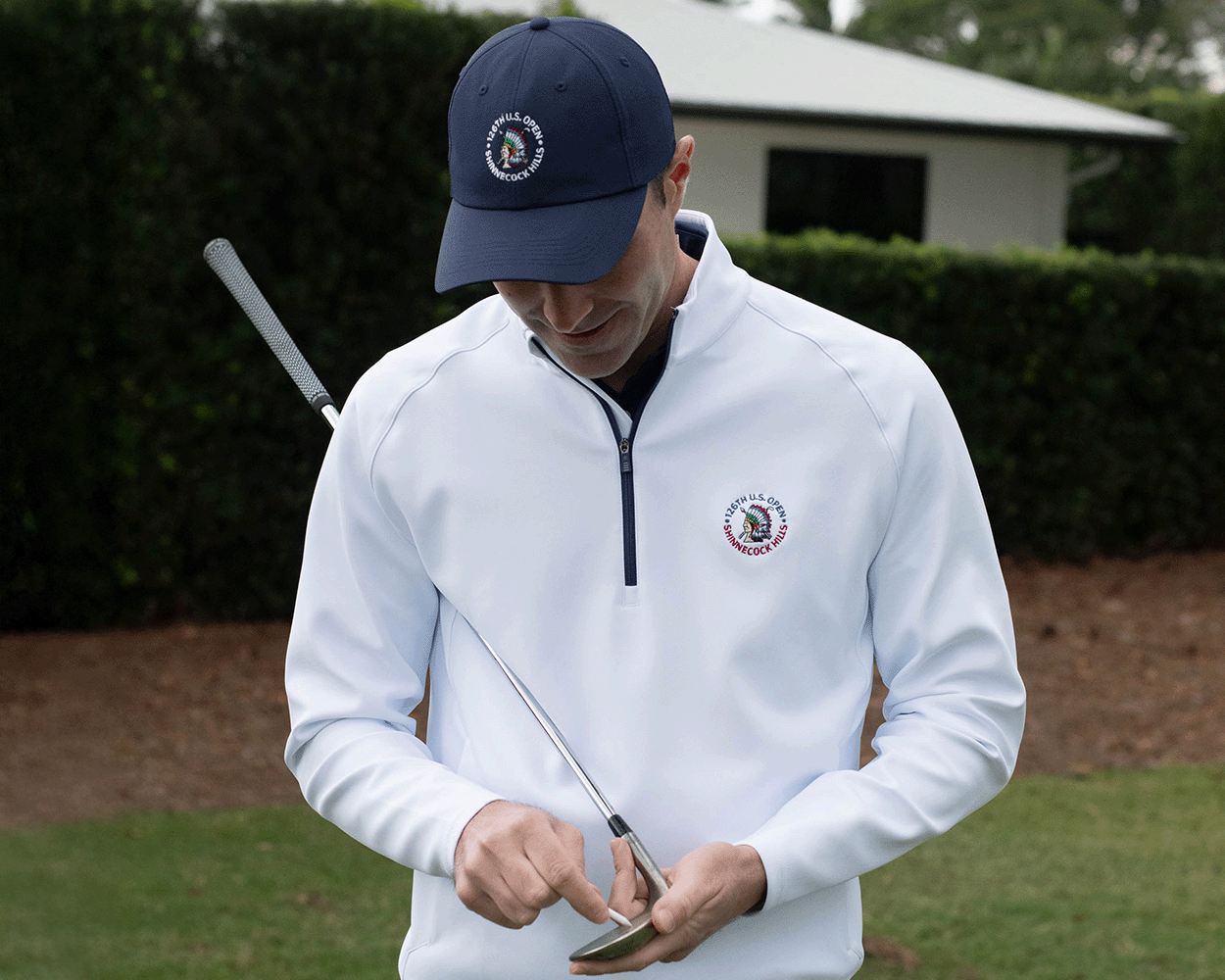 Man on golf course inspects putter, wearing 2026 U.S. Open Rodgers Pullover: White, featuring logo, UPF 50+ protection, and stretchy midweight fabric.