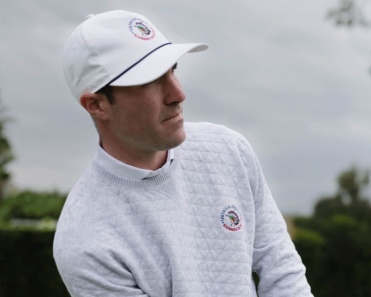 Man in 2026 U.S. Open Ward Sweater: Heathered Gray, featuring a quilt-textured pattern and logo, stands outdoors focused on his golf shot.