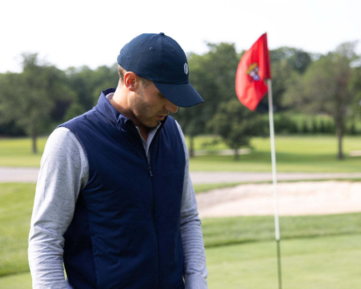 Man wearing navy hat and vest golf over a gray sweater on golf course with red flag in background.