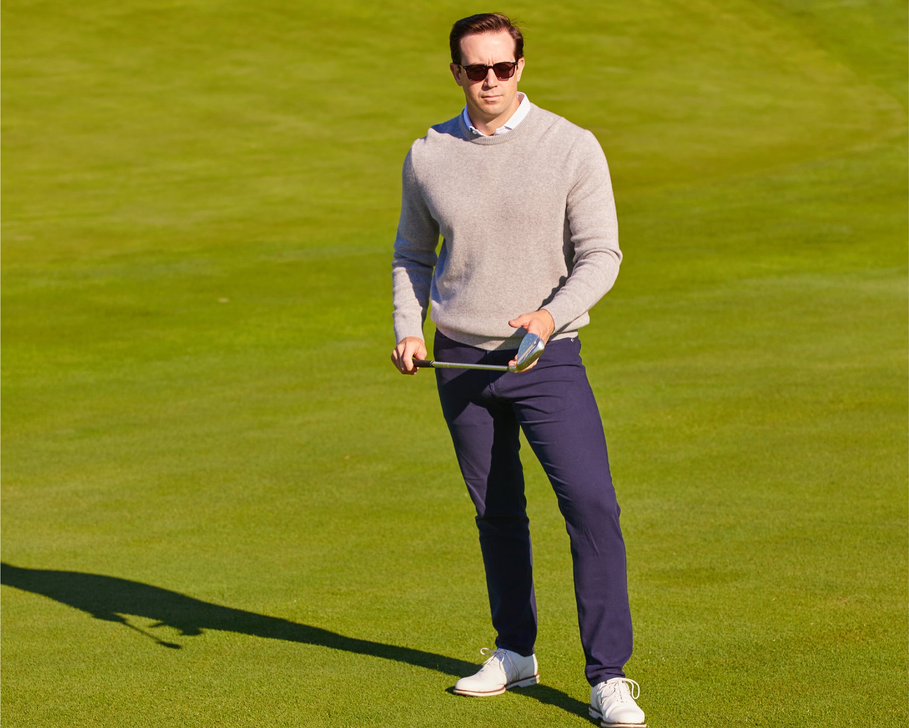 Man wearing Holderness and Bourne sweater gray stands on golf course holding golf club.