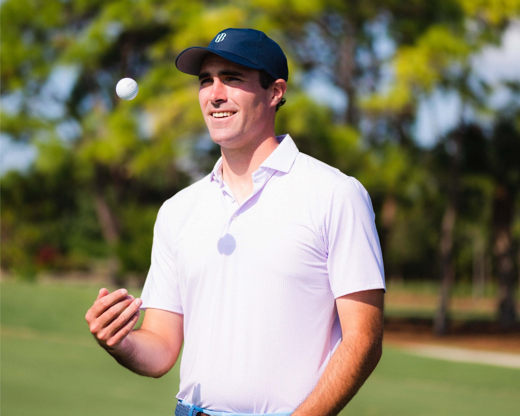 Male golfer wearing Holderness and Bourne navy promotional hat and white polo tosses golf ball in the air on golf course.