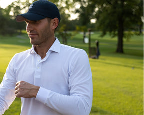 Male golfer wears Holderness and Bourne white long sleeve collared shirt and navy hat on golf course.
