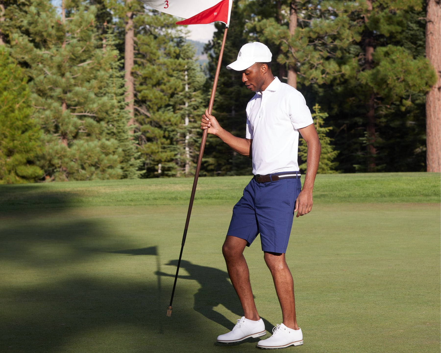 Male golfer wearing men white polo shirt holds red flag on golf course.