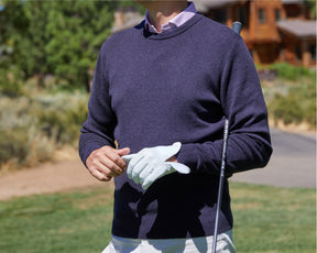 Male golfer wearing navy cashmere golf sweater holds club under armpit.