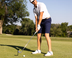 Male golfer wearing Holderness and Bourne white tailored shirt and navy shorts prepares to hit golf ball with club on golf course.