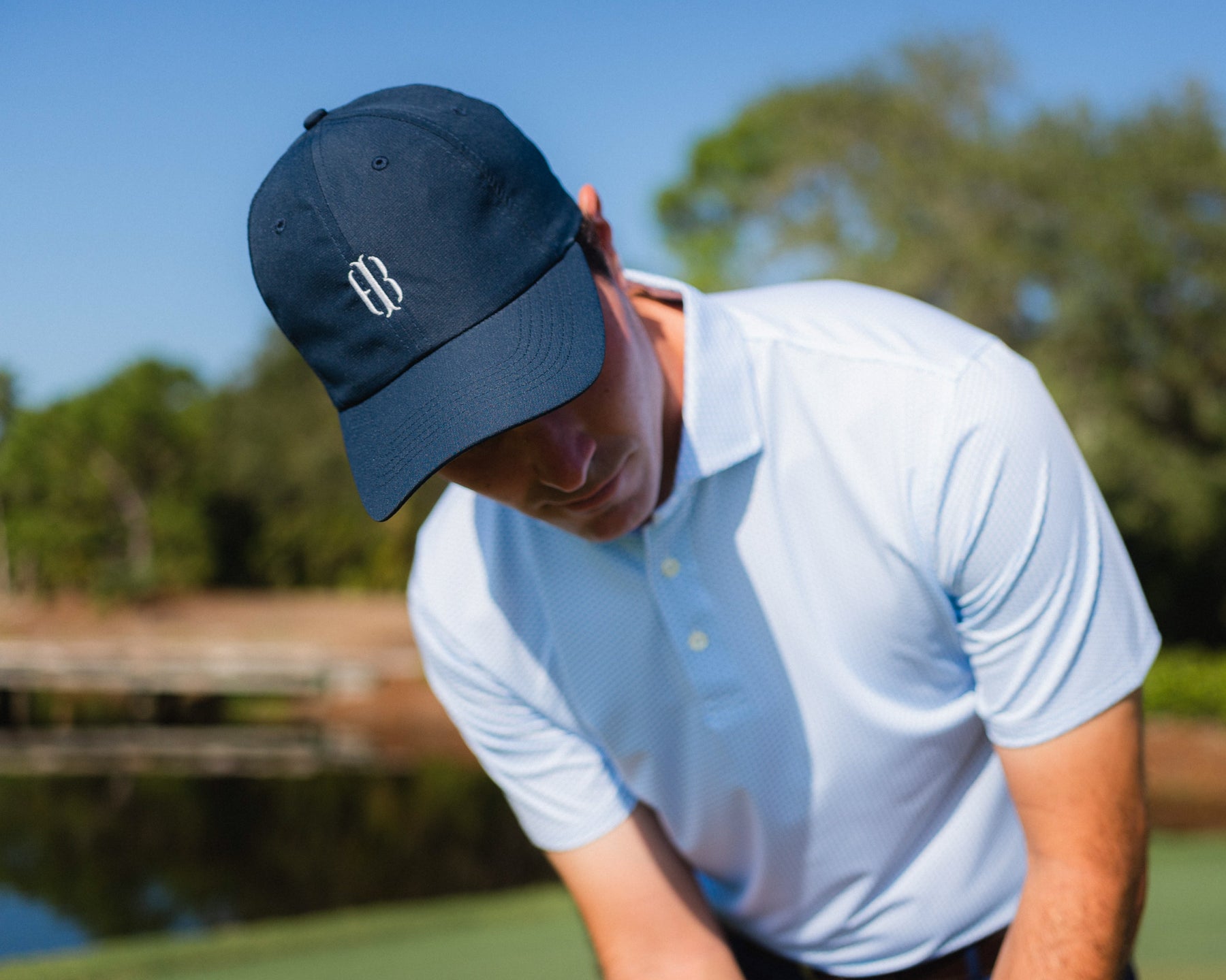 Male golfer wears Holderness and Bourne navy and white hat and white polo on golf course.