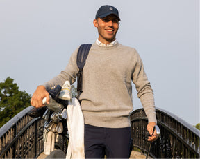 Man wearing heathered gray sweater and navy Holderness and Bourne hat hold golf club bag.